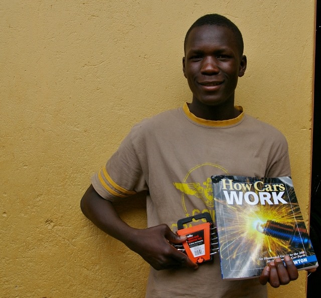 A boy holding tools and a magazine called 'How cars work'. He is now a mechanic thanks to S.A.L.V.E giving him an education!