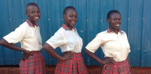 3 smiling girls stand next to each other wearing school uniform. Their right hands are on their hips and they are smiling at the camera.