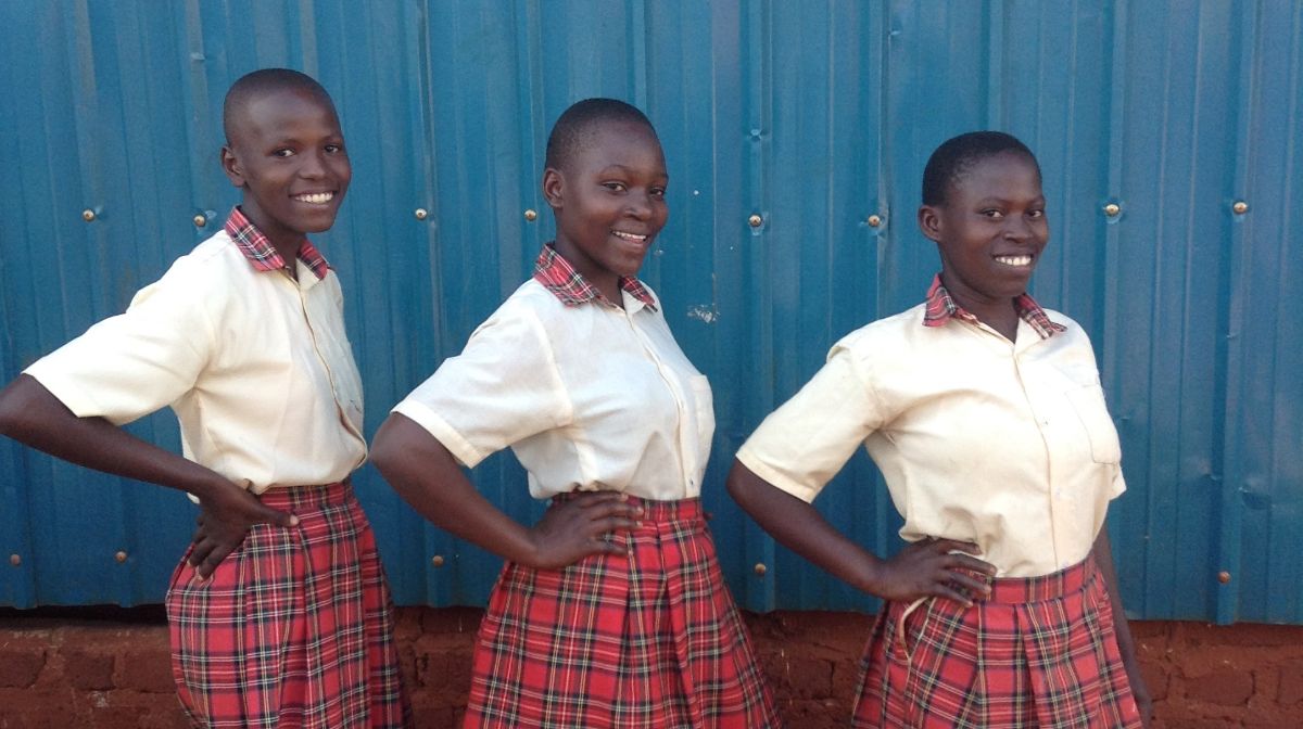 3 smiling girls stand next to each other wearing school uniform. Their right hands are on their hips and they are smiling at the camera.