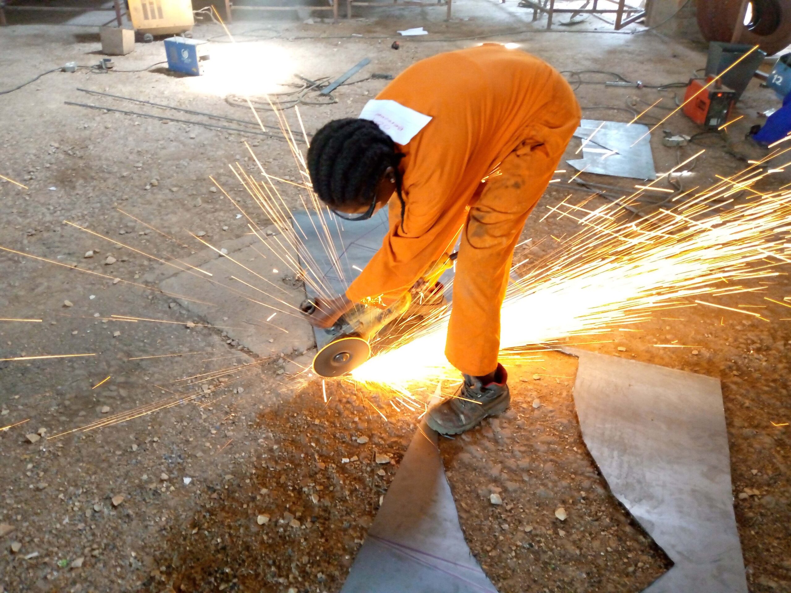 A girl wearing orange overalls, using an angle grinder on a piece of metal with sparks flying