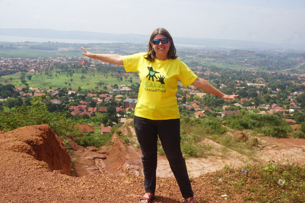 Nicola on a hill top in Uganda, with her arms out smiling. It is a sunny day and Nicola is wearing a SALVE t-shirt, looking happy.