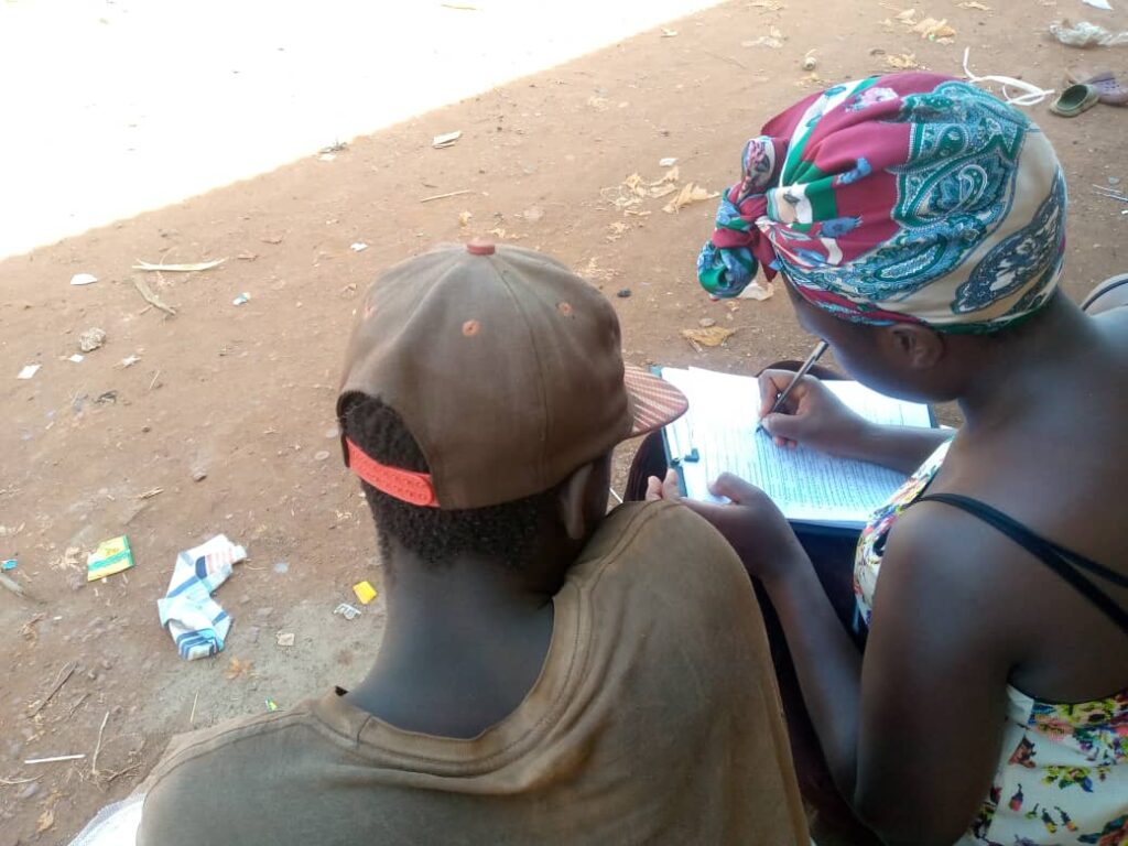 A female researcher is writing down the responses of a street connected boy. Both of their heads are bent towards the paper.