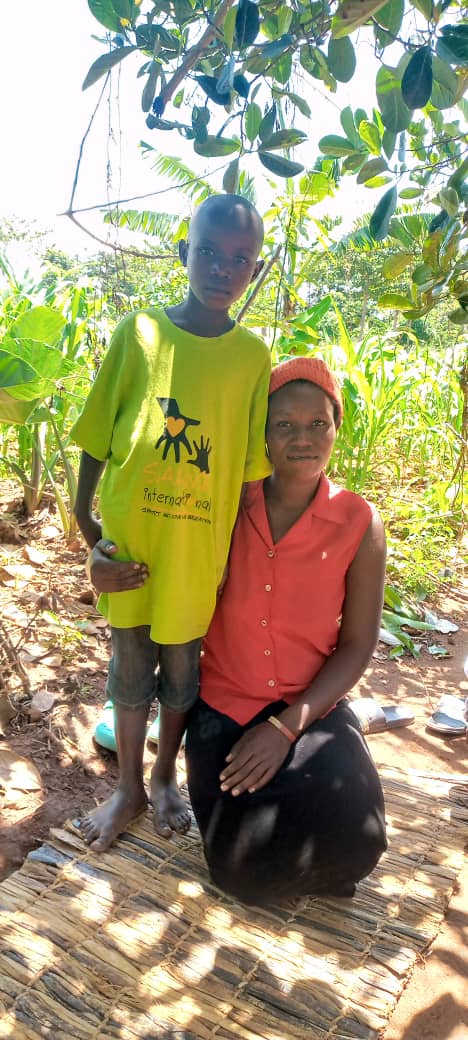 A young boy, stood next to his mother. His mother is kneeling on the floor and has her arm around him.